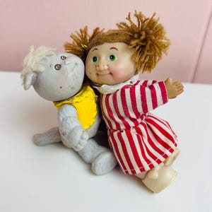 Peut inclure: Deux poupées vintage, l'une est un agneau gris portant un haut jaune, l'autre une fille dans une robe à rayures rouges et blanches. La poupée fille a des cheveux en fil marron et des yeux verts. Les poupées sont posées ensemble.