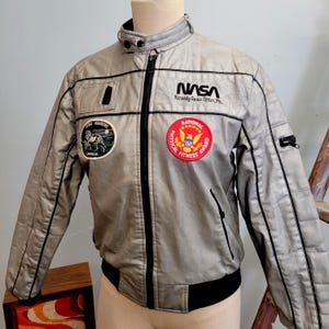 May include: Silver jacket with black trim and various patches, including the NASA logo and "Kennedy Space Center FL." It has a "National Physical Fitness Award" patch and a "Space Barons Canada" patch. The jacket has a zip closure and a stand-up collar.