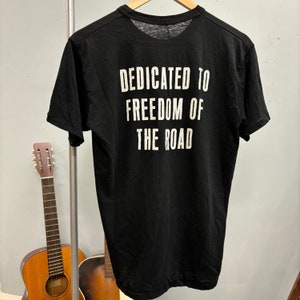 May include: Black t-shirt with the white text "DEDICATED TO FREEDOM OF THE ROAD" on the back. The shirt is hanging on a wooden hanger. Two acoustic guitars are in the background.