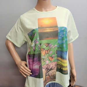 Puede incluir: Una camiseta de manga corta de color amarillo pálido con un gran collage gráfico central. El estampado incluye una puesta de sol sobre el océano, mariposas naranjas entre follaje verde y flores moradas, un árbol y motivos florales azules.