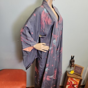 May include: A grey kimono with a floral pattern in shades of pink and blue. The kimono has wide sleeves and a V-neckline. The inside sleeve lining is a peach colour. The kimono is displayed on a mannequin.