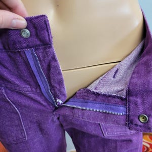 May include: Close-up of purple denim pants with a partially unzipped zipper. The pants have a button closure and a small pocket. The interior lining is a lighter shade of purple. The pants are displayed on a mannequin.