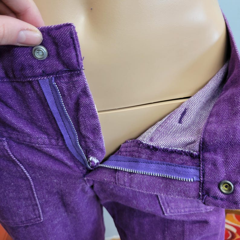 May include: Close-up of purple denim pants with a partially unzipped zipper. The pants have a button closure and a small pocket. The interior lining is a lighter shade of purple. The pants are displayed on a mannequin.