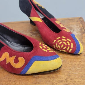 Puede incluir: Un par de zapatos de tacón de ante con base roja oscura y detalles vibrantes en amarillo, azul y naranja. Los zapatos presentan una puntera redondeada, un tacón bajo y un forro interior negro. Costuras amarillas crean un patrón circular.