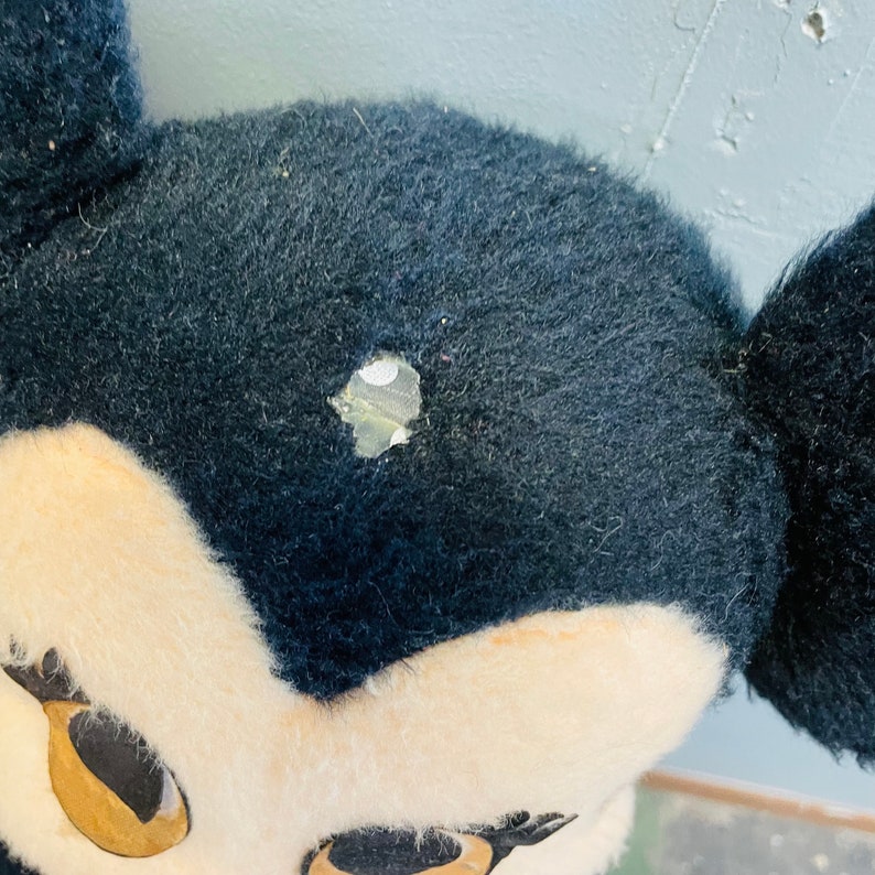May include: Close-up of a vintage plush toy head, featuring black fur and a cream-colored face. The toy has large, round eyes and a small hole in the head. The ears are black and stand upright. The toy appears to be well-loved and worn.