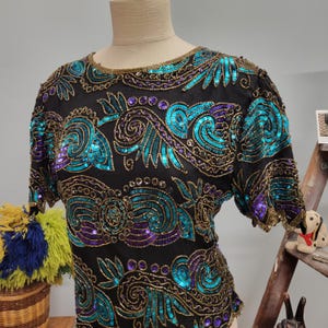 May include: Black short-sleeved top with teal, purple, and gold sequin embellishments. The sequins are arranged in a swirling, floral pattern across the bodice and sleeves. The neckline and sleeve edges are trimmed with gold beads. The top is displayed on a mannequin.