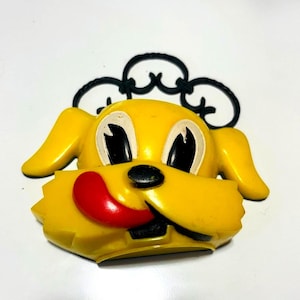 May include: A yellow plastic dog head wall ornament featuring large white and black eyes, a black nose, and a bright red tongue sticking out to the side. The piece includes a black decorative scrollwork hanger attached to the top of the head.