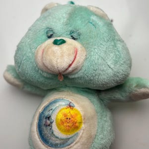 May include: A vintage teal plush bear with a smiling face, a green heart-shaped nose, and a tongue sticking out. The bear has a crescent moon and sun embroidered on its belly. A classic children's toy.