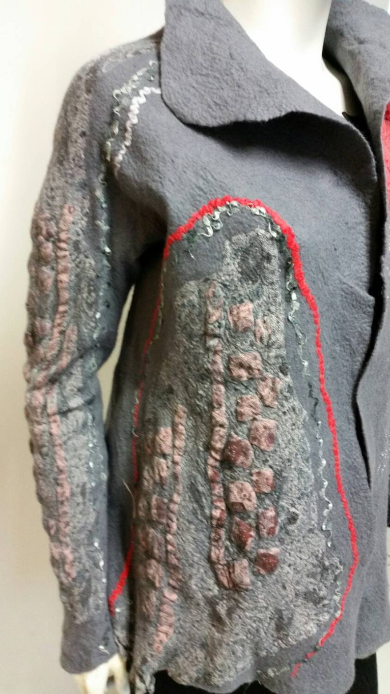 May include: A gray felted jacket with red and silver embroidery. The jacket has a collar and a button closure. The embroidery is in the shape of a flower.