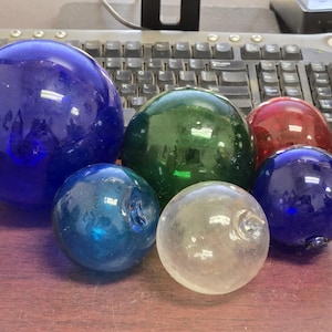 May include: A collection of six spherical glass floats in various sizes and colors, including a large cobalt blue sphere, a medium green sphere, a smaller red sphere, two smaller blue spheres, and one clear glass sphere, each with a small loop.