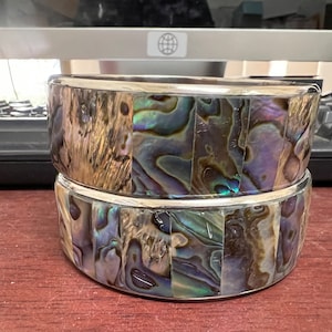 May include: Two stacked round silver and abalone shell boxes. The boxes are made of iridescent abalone shell tiles with a silver band around the top and bottom of each box.