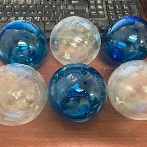 May include: Six glass spheres in varying shades of blue and clear. The spheres are arranged in two rows of three. The glass has a reflective quality, and the spheres appear to be decorative orbs.