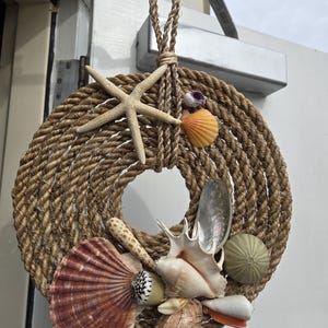 May include: A circular nautical wreath made of thick, twisted rope. Adorned with a variety of seashells, a starfish, and a sea urchin. The wreath is suspended by a rope loop, perfect for coastal decor.