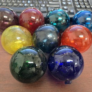 May include: A collection of nine spherical glass floats arranged in a triangular formation. The smooth, translucent orbs feature a variety of vibrant colors including deep purple, bright red, teal, light blue, yellow, black, orange, green, and navy.