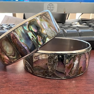 May include: Two silver-toned, wide bangle bracelets. The bracelets feature iridescent abalone shell inlays, showcasing a spectrum of colors including blues, greens, and purples. The shells are set in a rectangular pattern. The bracelets are stacked on top of each other.