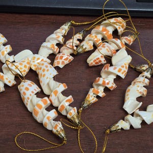 May include: Several spiral-shaped seashell ornaments with orange and white patterns. Each ornament has a gold-colored string for hanging. The seashells are arranged on a dark brown surface, suggesting a coastal or nautical theme.