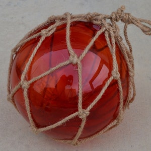 May include: A round, red glass float, encased in a beige rope netting. The float has a reflective surface, and the rope is knotted at the top. This decorative item is suitable for nautical-themed decor.