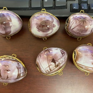 May include: Six decorative shell-shaped trinket boxes with gold-colored metal trim. Each shell features a different coastal scene in shades of purple and white, including mermaids, turtles, and sailboats. The shells are displayed on a brown surface.