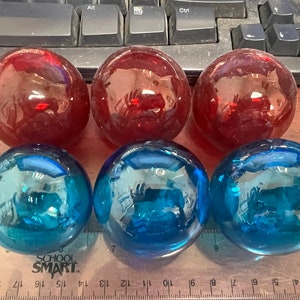 May include: Six glass spheres, three in a deep red and three in a vibrant blue. The spheres are arranged in two rows of three, with a ruler visible at the bottom. The spheres are reflective and appear to be solid.