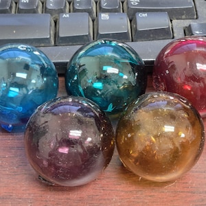 May include: Five colorful glass spheres in shades of blue, green, red, purple, and amber. The spheres are transparent and reflect light, creating a glossy appearance. The objects are arranged on a wooden surface, with a keyboard in the background.