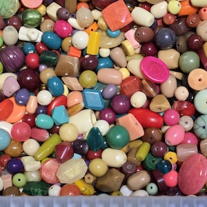 May include: A close-up shot of a collection of colorful beads in various shapes and sizes. The beads are in shades of pink, red, green, blue, yellow, and brown. Some beads are round, while others are rectangular or faceted.