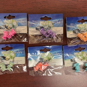May include: Six pairs of shell jewelry earrings in various colors, including pink, purple, orange, green, red-orange, and turquoise. Each pair is packaged on a blue and white card that reads "Genuine Shell Jewelry."