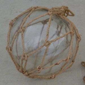 May include: A clear glass buoy with a natural brown rope net. The buoy is round and has a knotted rope handle.