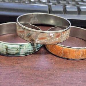 May include: Three silver-toned bangles with inlaid decorative elements. One bangle features a mosaic of white and tan pieces, another has a green and white pattern, and the third has an orange and tan design. The bangles are displayed on a brown surface.