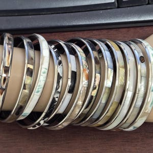 May include: A collection of silver bangles with inlaid mother-of-pearl and other decorative elements. The bracelets are in various colors, including white, black, and iridescent hues. The bangles are stacked on a wrist.