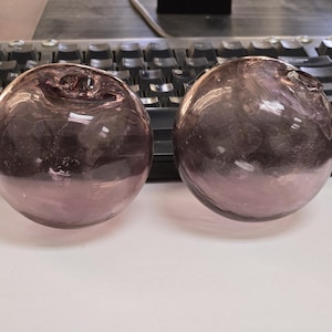 May include: Two round, purple glass objects with a slightly flattened top. The glass has a translucent quality, with a darker shade at the base. The objects are sitting on a white surface with a keyboard in the background.