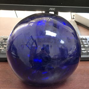 May include: A large, spherical, deep blue glass ball. The surface reflects light, creating bright highlights and distorted reflections of the surroundings. The object is smooth and appears to be made of a solid, transparent material.