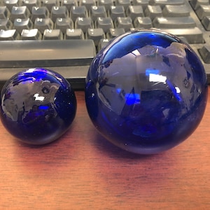 May include: Two blue glass spheres, one larger than the other, on a wooden surface.