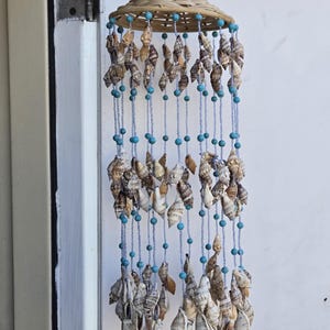 May include: A hanging wind chime featuring a woven light brown bamboo top structure. Multiple strands of light blue beads and small, variegated brown and white seashells hang from the base, creating a tiered, cascading arrangement of natural shells.