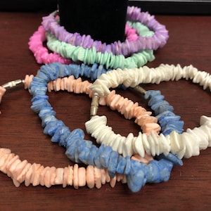 May include: A set of five shell bracelets in various pastel colors, including pink, light green, lavender, white, and blue. The bracelets are made of small, flat pieces of shell that are strung together on a silver chain.