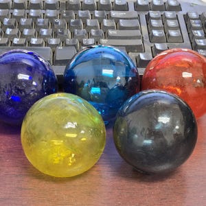 May include: A collection of five smooth, spherical glass floats in various colors including deep blue, bright blue, translucent red, yellow, and black. These round glass orbs are arranged in a cluster, each featuring a polished, reflective surface that catches light, creating distinct highlights across their curved, circular forms.