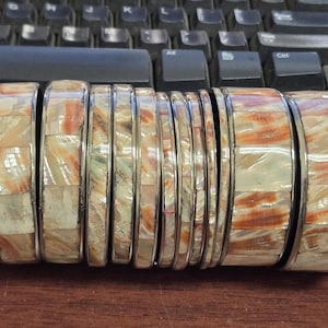 May include: A set of eight bangle bracelets featuring a metallic frame and inlaid rectangular tiles of iridescent shell material. The tiles display a marbled pattern with shades of cream, beige, and reddish-brown, arranged in a repeating grid.