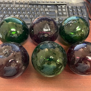 May include: Six glass floats, three green and three purple, are arranged on a wooden surface. The floats are round and have a smooth, shiny finish.