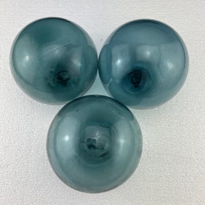 May include: Three teal blue glass fishing floats. The floats are round and have a smooth, shiny surface.