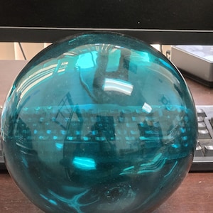 May include: A large, spherical, teal-colored glass ball. The surface reflects the surrounding environment, including a keyboard and monitor. The ball has a smooth, glossy finish and appears to be made of thick glass.