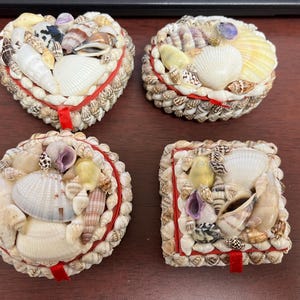 May include: Four decorative boxes made of seashells. The boxes are in the shapes of a heart, a square, and two circles. The boxes are all decorated with a red ribbon.