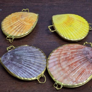 May include: Four scallop shell pendants with gold-toned metal edges. Each shell features a unique color: one orange, one yellow, one purple, and one pink. Each pendant includes small gold metal loops attached to the sides for hanging purposes.