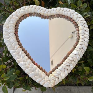 May include: Heart-shaped mirror with a frame made of white seashells. The shells are arranged in a layered pattern around the mirror's edge. The mirror is surrounded by green foliage.