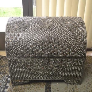 May include: A silver metal chest with a decorative pattern of small circles and swirls. The chest has a hinged lid and is closed.