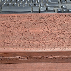 May include: A rectangular wooden box with intricate floral carvings. The box is a warm brown color and features a detailed pattern of flowers and leaves across the lid. The sides of the box also have carved details.