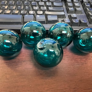 May include: Five teal blue glass ornaments, each with a slightly different shape and texture, are arranged on a wooden surface.