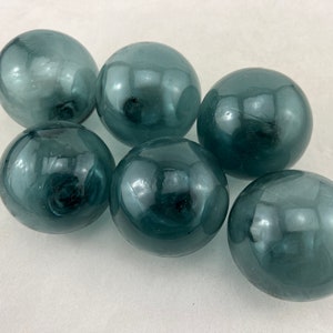May include: Six teal-colored glass spheres arranged on a white surface. The spheres have a glossy finish and reflect light, creating highlights. These decorative orbs could be used for home decor or crafts.