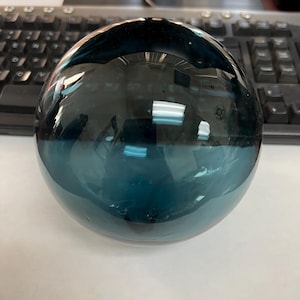 May include: A large, round, teal blue glass orb. The orb is smooth and reflective, and it appears to be made of blown glass.