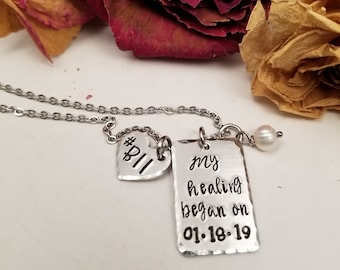 May include: Silver necklace with a heart charm that says "#BII" and a rectangular charm that says "my healing began on 01.18.19".