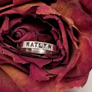May include: A silver ring with the name "KATLYN" engraved on it. The ring is resting on a dried red rose.