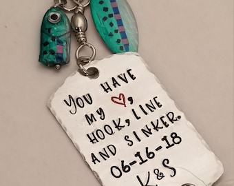May include: A silver-colored keychain with a fish-shaped charm, a teal and blue lure, and a rectangular tag. The tag reads "You have my hook, line, and sinker. 06-16-18 K&S" in black lettering. A small feather is attached.